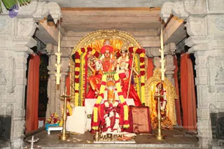 A photograph of a temple shrine with a golden arch backdrop and carved stone pillars. Two deity murtis are seated under the arch, one in red with a red headdress and the other in a crown-like headdress, both decorated with thick garlands of yellow and red flowers. A red-and-gold cloth hangs behind them and a smaller golden shrine stands to the right. Brass oil lamps, a kalasha and flower offerings sit on the stone platform in front. No people are visible in the scene.