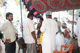 Outdoor relief camp under a red and green embroidered canopy with a purple fringed edge, beside palm trees. A man in a white shirt and dark trousers stands at left with hand on hip. A man in a striped shirt and khaki trousers talks with a woman in a brown sari. A man in white robes and a white head cap stands center. Another man in a white cap sits on an orange plastic chair, and a shirtless man with a white cloth wrap sits beside him. Sacks and vehicles are visible behind, and a man in white stands at far right.