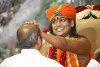 A photograph of a smiling man in an orange turban with a gold brooch and red bindi, wearing an orange saffron garment with gold borders and a red-and-white flower garland with rudraksha beads. He rests his hand on the head of a balding man seen from behind in a white shirt, in a ceremonial setting with a floral backdrop of pink and white blossoms and a garlanded golden seat. Swamiji (probable) appearance cues: orange turban and saffron clothing with rudraksha malas, seated in a ceremonial blessing role. Two people visible.