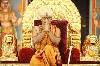 A seated man in saffron-orange robes with a cream turban and red forehead mark sits with palms pressed together in prayer on an ornate gold throne. The throne has a gold backrest with lion-form arms and red velvet cushioning, flanked by two golden lion statues. A small microphone is clipped near his face. Behind him, a painted deity figure with multiple arms and gold ornaments is visible. He wears layered beaded necklaces and bangles. Swamiji (probable) is shown with long swept hair absent, but the saffron robes, turban, multiple rudraksha-style malas and ceremonial gold throne setting mark him as a.