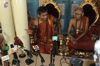 A press conference beside golden thrones with carved gilt arms and red cushions, behind a table crowded with microphones bearing Tamil news channel logos. A young man in saffron-orange robes, rudraksha malas, a beaded headband and a red-orange kumkum forehead mark stands at the table pointing with his right hand, an armband on his upper arm. Seated to his right on the throne is an elderly bald man in saffron robes with white vibhuti markings, heavy rudraksha malas and a gold ornament, his hands resting together in his lap. Blue carved doors and a cream pillar form the background. Two people are visible at the left edge.