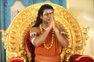 A man seated on an ornate gold throne with a carved, scrolled backrest and red velvet cushions, pressing his palms together in anjali namaskar at chest height. He wears an orange-saffron cloth wrap over his shoulders, a multi-strand necklace of large gold beads, rudraksha-style brown beaded bands on both upper arms, and a white vertical tilak with a red dot on his forehead. His long dark hair falls past his shoulders. Behind him are pale blue and cream walls with decorative painted panels. The image carries a faint copyright overlay in the lower right. Appearance-based label: Swamiji (probable), seated in a throne-like setting with long hair, saffron clothing, gold beaded ornaments and rudraksha-style beads.