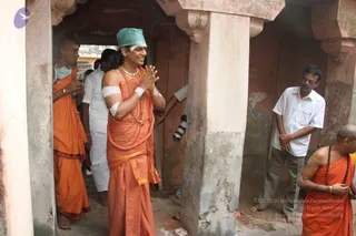 A man in saffron-orange robes stands smiling with palms pressed together in a namaste gesture inside a worn stone temple corridor with pink-washed walls and a thick pillar. He wears a teal cloth cap, white forehead markings, a bandage on his upper arm, and long rudraksha malas with a white ceremonial necklace. Around him, orange-robed sannyasis and men in white shirts and trousers stand near the doorway, and a bald monk in orange with beads is at the right edge. A man in a white shirt and glasses watches nearby.