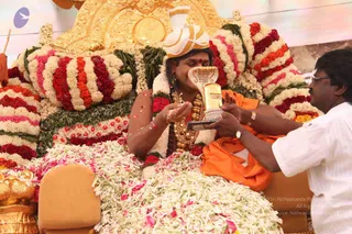 A seated figure in an orange-saffron robe and a white and gold turban sits on an ornate golden throne heavily decorated with red, white, pink and orange flower garlands. He wears layered gold ceremonial necklaces, a rudraksha-style bead mala, and a red tilak on his forehead, and his eyes are closed as he receives a gold ceremonial object with a cobra-hood top. A man in a white shirt and glasses, standing at the right, holds the gold vessel toward him while white and pink petals scatter. White jasmine garlands cover the floor in front of the throne, with a gold pot visible at the lower left. The scene suggests a devotional offering at a satsang.