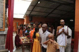A photograph of a covered courtyard at a temple-style ashram with orange walls and a corrugated roof. A shaved-headed young man in a white dhoti with a gold border and a rudraksha mala stands at right with his palms pressed together, sacred ash marks on his forehead. Around him, several shaved-headed boys and girls in orange, red and yellow clothing gather near a small wooden table holding brass vessels and bowls. A woman in a light top and other people move through the space. Carved stone pillars and a red curtain frame the left side.