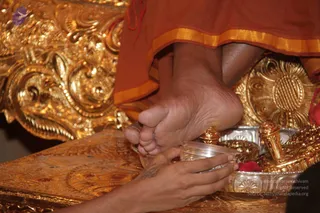 Close-up photograph of a bare foot in an orange-saffron garment with a gold-bordered hem resting on a silver tray. A hand with a henna-like tattoo holds a clear bowl and sprinkles or pours liquid over the foot, a padapuja style foot-washing. Ornate gold-embossed throne or peetha fabric and gold ritual vessels surround the scene, with a red flower petal on the tray. Only the feet and hand of people are visible; no face is shown.