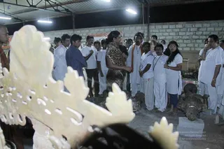 A photograph taken indoors at night under bright ceiling lights in a open-sided workshop with a corrugated roof and a cinder-block wall. A crowd of roughly two dozen people in white clothing stands in a semicircle around a stone sculpture workspace. A woman in a leopard-print top stands at the centre, gesturing as she speaks. A man in a white kurta with an orange shawl and a rudraksha-style necklace stands among the group. Foreground includes a large cream carved stone ornament with openwork foliage and a dark bronze or metal figure on a plinth to the right. Some onlookers wear white tees and lanyards. The scene looks like a guided visit or demonstration around carved and cast religious artwork.