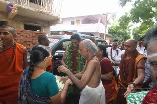 A street scene during a house visit in Tamil Nadu. A bare-chested, shaved-headed man in a white dhoti with red tilak and rudraksha mala stands centre, receiving a green leaf garland from a man in a red-maroon robe. A woman in a teal blouse and blue sari holds a basket of white flowers and reaches toward him. On the right, a shaved-headed monk in saffron-orange robes and a red-brown shawl with rudraksha beads stands near others in white and red-maroon. On the left, a young saffron-robed monk with rudraksha mala stands at the edge. Behind them a dark vehicle with a red-and-white sign is parked before a brick wall and a pink building.