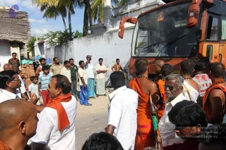 A street scene in a village beside a whitewashed compound wall and a thatched hut, with palm trees behind. An orange-and-red bus fills the right side of the frame. In the foreground, a group of devotees stands in the road: a shaved-headed man in an orange cloth with a rudraksha mala and gold chain at the centre, another shaved-headed man in orange at the right, and men in white shirts and white dhotis, one older man with glasses and a white shirt holding a green-handled staff. Several local villagers in lungis and shirts watch from the left behind a low wall, and a man in a white shirt and orange shawl stands at the left edge. No a person candidate is visibly identifiable, so the figures are described by role only.