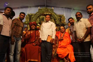 Two seated saffron-clad figures sit on an ornate throne-like chair in front of a green and gold temple-style arch with a printed deity image and flower garlands. On the left, a figure in a deep red-orange turban and wrap wears rudraksha beads and smiles while raising a hand. On the right, a figure in orange robes and a dark shawl with a white beaded necklace sits with palms together. A man in a white shirt and dark trousers stands between them holding a small book, flanked by men in casual shirts. The scene is a photograph of a seated ceremonial gathering.