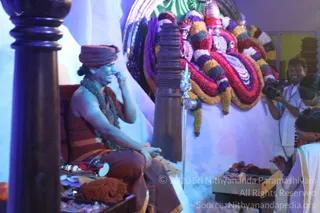 A photograph of a satsang setting with a seated figure on an ornate red-cushioned throne at left. The figure wears an orange-brown turban, a long dark rudraksha mala draped over the shoulders, and an orange-and-brown robe, and holds one hand near the face in a gesture. Beside the throne is a tall dark wooden pillar and a metal trident leaning against a stand. Behind is a large deity murti or decorated backdrop garlanded with bands of red, purple, yellow and white flowers, with a silver-toned crown-like element above. A cameraman in white shirt films at right, and a white-clad person with a dark head covering sits in the foreground near a low table with fruit and offerings. Stacked cushions and brass items lie in the lower left.