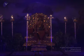 A dimly lit, blue-violet stage set shows a seated figure on an ornate golden throne with a large golden sunburst-style backrest, framed by tall silver trident staffs with decorative finials and dark foliage on both sides. The figure wears orange-red draped cloth and a beaded necklace, with long hair visible, and sits on a golden dais above a patterned mat. A dark cloud-like backdrop and an umbrella-like canopy shape rise behind the throne. a person: the seated figure has long hair, an orange garment and beaded ornaments, consistent with the page's Kalabhairava darshan setting, but the face is too dim to confirm. The image appears to be a tableau or statue-like arrangement rather than clearly live people.