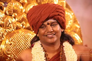 Close-up photograph of a smiling man seated before an ornate gold repoussé backdrop with scalloped patterns. He wears a rust-orange turban, a dark red-brown dot on his forehead, white jasmine garlands, and rudraksha beads with gold necklaces and bangles on his bare shoulders. Swamiji (probable): the long dark hair falling behind the shoulders, the orange turban, rudraksha malas and ceremonial gold cues align with the page's note that Swamiji is at this event, so this is a probable appearance-based candidate, presented as a seated devotee-facing figure in a photo.