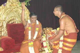 A seated figure on an ornate gold-carved throne with a red upholstered seat raises one arm and holds a green leaf aloft while smiling. The seated figure wears an orange-saffron robe with a multicoloured garland of white flowers and red, yellow and green blooms, a mala of beads, and a red bindi. Beside the throne, a standing man in a saffron and red-orange dhoti and shawl, with shaved head, glasses and a beaded bracelet, offers a garland and holds a garland-decorated object. A dark maroon curtain forms the backdrop. Swamiji (probable): long dark hair swept back, orange robe, rudraksha mala and garland on the seated figure, seated on a throne.