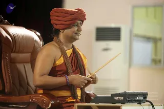 A man in a rust-orange turban and rust-orange and yellow sash sits in a large brown leather armchair, holding a wooden drumstick in his right hand. He wears a long rudraksha mala with a gold-toned element around his neck and a red and blue beaded bracelet on his wrist. Beside him is a black DTX electronic drum pad on a low wooden table, and a white wall-mounted air conditioning unit stands behind him near a window. A small blue-and-white logo appears in the top left corner. The scene suggests a live session or teaching event in an indoor hall.