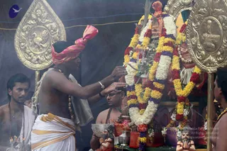 A ritual scene in a smoky tent-like setting. A bare-chested priest in a pink and gold turban and white dhoti with a gold border stands at left, reaching toward a deity murti draped with garlands of white, yellow, red and purple flowers. Golden ceremonial shield-shaped backdrops frame the altar. Another shirtless man with a rudraksha mala, and a man holding a brass bowl, stand beside the altar, with carved figures and brass vessels in front. A mild haze of smoke or steam hangs in the air. Appears to be a homa or puja ritual.
