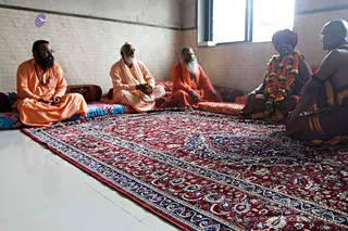 Five men sit cross-legged on cushions and bolsters around a large red and blue patterned carpet in a plain room with white tiled walls and a window. On the left, a bearded man in saffron-orange robes sits facing right. Next is an older white-bearded man in cream-white robes with a light shawl. Beside him a white-bearded sadhu wears orange-saffron robes. On the right, a sadhu in maroon-brown cloth wears a dark cap and a garland of orange and white flowers, and at the far right a bald man in maroon-and-gold-bordered dhoti sits in profile. Their hands rest in their laps, and they appear to be in a seated discussion.