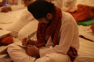 A bearded man in a black turban and white kurta sits cross-legged on a white floor covering, bent over a notepad and writing with a silver pen. He wears a red shawl printed with dark script. Behind him other seated people in white and orange-red cloth are visible, and a green and red mat lies to the right. The setting appears to be a satsang gathering where participants take notes.