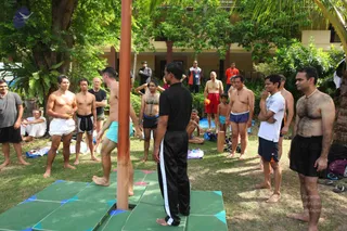 Outdoor photo in a garden courtyard with a tall wooden pole standing upright in the foreground on a green-and-teal mat platform. A shirtless man in light blue swim trunks stands on the platform beside the pole, while a man in a black athletic top with white side stripes stands facing him. Around them, a loose group of mostly shirtless men in shorts and swim trunks watch, some with arms crossed, one in a white shirt and dark shorts, and a man in red shorts near a building. A young boy in blue shorts stands among them. Palm and tropical trees, a two-storey cream building with columns, and folded clothes and bags on the grass fill the background.