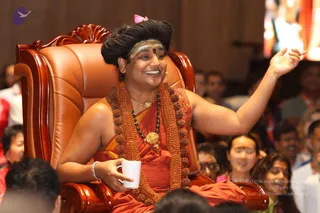 A photograph of a smiling person seated on an ornate carved wooden throne with brown leather cushions, one arm raised outward and a white mug in the other hand. The seated figure has long dark hair swept up, a white and orange tilak on the forehead with a red dot, an orange-red robe, a long orange shawl draped over the shoulders and a dense cord of rudraksha beads, a green bead mala, and a gold pendant necklace. A pink bow is visible in the hair. Behind and around the throne, an audience of mostly seated people, some wearing red and orange lanyards, watches attentively. Appearance-based identification is probable for a person based on the visible cues of swept hair, saffron-orange clothing, rudraksha mala and ceremonial gold necklace, seated on a throne.