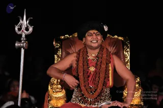 A man seated in a gilded, brown-upholstered throne-style chair against a dark background, smiling. He wears a thick multi-strand rudraksha mala of brown beads, gold necklaces with a large gold pendant, a red-and-gold wrap, a leopard-print garment, a red bindi and a red-and-gold bracelet, with dark hair swept up and a silver jewel pin. A silver trident stands to the left, and gold lion-head carvings decorate the chair arms. Audience members are faintly visible at the edges.