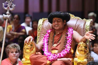 A man seated on an ornate gold-trimmed brown leather throne with carved lion-head arms sits in a saffron-orange robe with a gold border. He wears a long rudraksha mala, a green bead necklace, a ceremonial gold pendant and a magenta orchid garland, with dark hair swept up, a red bindi and a head-mounted microphone. He is eyes closed, speaking, with one hand resting on a lion armrest and the other open in a gesture. A tall silver trident stands at left. Behind him a seated audience of devotees in saris and kurtas, one woman in orange in the foreground, fills a hall with purple and gold drapery. Watermarked archive photo of a discourse.