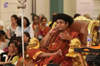 A photograph of a long-haired man in saffron-orange robes with a rudraksha mala and a large rudraksha necklace, seated cross-legged on an ornate gold-and-brown throne with carved lion-head armrests. He wears a head microphone, a red forehead mark and bangles, and holds his hand near his face while speaking. Seated audience members in orange and white clothing, some with lanyards, fill the hall behind him. A silver staff stands at left and a water bottle sits on a side table at right. Swamiji (probable): long swept-back hair, saffron robes, rudraksha malas and ornate gold throne are the cues, and this matches the page's note that he is present at the event.