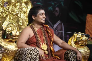 A photograph of a seated man on an ornate golden throne with lion-head armrests, speaking into a microphone on a stand. He wears a rust-orange robe draped over one shoulder with a gold-and-orange striped shawl, multiple rudraksha mala beads, a heavy gold ceremonial necklace with a pendant, and a gold ring. His forehead bears white vibhuti and a red kumkum dot, and he has long swept-back dark hair. His leopard-print cloth covers his lap. Behind him, a dark backdrop shows a painted or printed figure of a bearded sannyasi in white. Probable Swamiji, seated in discourse, judged from the distinctive regalia of rudraksha malas, orange robe, gold necklace and long hair.