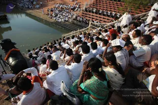A large crowd of devotees sits in tiers on stone steps beside a rectangular temple tank of still water at dusk. Most wear white ritual clothing; several women wear green and patterned saris and some men wear white dhotis and caps. Many have hands raised in namaskar or folded in prayer, and some are seated with eyes closed. On the left, a black stone Nandi bull with a marigold garland lies at the water's edge. Brick and stone terraces and a metal railing line the bank behind the seated crowd, with more people visible on upper steps. The scene suggests a collective puja or darshan at an outdoor tank.