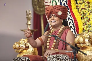 A seated man in a rust-red turban with a jewelled brooch and a red tilak on his forehead gestures with his raised right hand while speaking into a microphone on a stand. He wears a rust-red robe, a long necklace of large gold beads over a brown rudraksha-style mala, and red beaded bracelets. He sits on a throne with golden lion-head armrests and a leopard-print cushion, backed by a garland of yellow and red flowers and a patterned cloth. A golden oil lamp stands at the left.