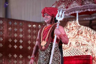 A smiling man stands on a stage holding a silver trident in his right hand. He wears a red turban, a red shawl over a bare upper body, a brown rudraksha mala, a gold floral garland and a gold pendant, and gold bangles on his wrists. He wears a leopard-print lower garment and large gold earrings. Behind him is an ornate gold throne with carved details and a red velvet cushion, under a red and gold embroidered canopy with tassels. A red patterned wall panel forms the backdrop.