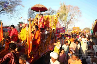 A festival procession at a Kumbh Mela-style event on a dusty roadside under bare trees. A decorated float is draped in marigold garlands and crowned by a red and gold umbrella. Several men in saffron and orange robes ride on the float, one holding a long saffron flag on a pole; some wear red shawls and others are shirtless with orange cloth. A dense crowd of men in white dhotis and turbans, women in saris and a young girl in pink surround the float. Other decorated floats with orange and gold decorations follow in the background. A banner with red lettering is visible among the crowd.