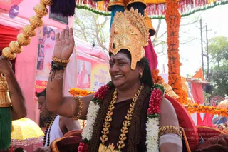 A smiling man rides in a procession under an orange marigold-garlanded canopy, raising his right hand in greeting. He wears an ornate gold crown with a red gem, gold armbands and bangles, a dark maroon robe, and multiple garlands of white jasmine and red flowers, with gold necklaces and a rudraksha-style bead mala. A red tilak mark is on his forehead. Behind him stand a man in white and another attendant with a gold-topped staff and a yellow-fringed umbrella on the left. Pink and red tents and a hoarding with portraits and Hindi text line the street, with crowds and banners in the background.