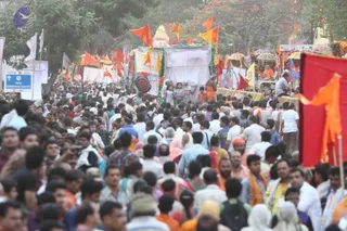 A dense outdoor procession crowd moves along a tree-lined road under a hazy sky, with many people in white, cream and orange clothing. Saffron and orange flags rise above the throng, and a white-draped float or ratha with decorated pillars and umbrellas carries a few people in saffron robes near its top. A large red banner with orange pennants stands in the right foreground, and a blue sign reading drinking water appears at the left. Most people in the crowd are walking, and many are close-packed and partly obscured.