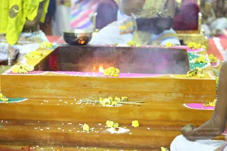 Close view of a wooden fire pit (homa kunda) lined with pink and green powder and scattered with yellow marigold flowers and white grains, with a small flame and rising smoke at its centre. A brass bowl with a lit wick sits behind it, and partially visible seated people in white and yellow-green clothes surround the altar. Only hands and limbs of a seated person appear at the right edge.