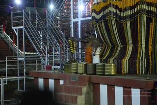 Night photograph of a temple-style stage at an outdoor ritual venue. Two men stand at the platform beside a small metal deity murti draped with a yellow and orange marigold garland. The man in the centre wears bare-chested with an orange dhoti and gestures toward the murti with his hands raised; the man beside him wears a white dhoti and shawl and stands close. Five polished brass pots or kalashas sit in a row in front of the murti on the red-and-white striped platform. Behind them a large wall is covered in layered garlands of marigold, white and maroon flowers. Metal scaffolding with stairs and bright floodlights frames the scene, with rows of seating and red flags in the background.