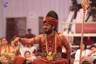 A photograph of a seated man in a rust-red turban with a jewelled ornament and rust-red robes, wearing a leopard-print waist cloth, heavy gold necklaces and rudraksha malas, and rust-red bracelets and armlet. He rests one hand on a brass animal-headed armrest and holds a tall gold staff with a trident-like top to his right. Behind him an audience in white and pale clothing sits on a stage with white drapery, a red banner and camera equipment. Swamiji (probable): the rust-red and saffron clothing, turban and long hair with gold necklaces are the cues, seated as the speaker.