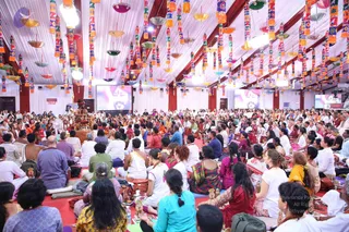 A large indoor satsang hall with white draped ceiling and hanging colourful decorative lanterns in pink, orange, yellow and green. A large crowd of devotees sits on red floor mats facing a raised seat at the front, where a figure in orange-saffron clothing sits on a platform. Large screens on the walls show a close-up face. Many attendees wear white, pink, saffron and green clothing; some hold water bottles and bags. The atmosphere is a seated assembly with a central speaker.