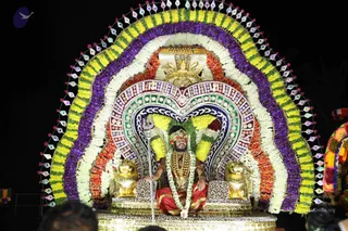 Night procession photograph of a man seated on an ornate golden throne (peetha) inside a large arched floral canopy of marigolds and white garlands in purple, yellow, orange and white, with a decorative multicoloured panel behind him and a crown-like golden finial above. Two golden lion figures flank the throne. He wears a red-and-gold dhoti, a long rudraksha mala and a flower garland, with a red headband and a trident (trishul) held upright beside him. Devotees are partly visible at the lower edge of the frame. a person (dressed in saffron-red with rudraksha and ornate headwear) seated on the throne as the honoured figure of the procession.