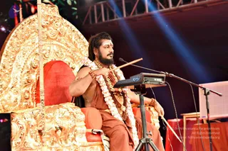 A man seated on an ornate golden throne with a red cushioned seat, speaking into a microphone on a stand with a DTX-labelled electronic device before him. He wears a rust-orange robe, a white-flowered garland, layered gold-coloured necklaces and beaded bangles, with a bindi and horizontal white tilak on his forehead, and holds a slender wooden stick in one hand. Blue stage lights and a metal truss frame the stage behind him, with a pink-draped table at the side. Swamiji (probable): long, swept-back hair, a crown-like ornate gold throne setting, saffron-orange clothing, rudraksha-style beads and ceremonial gold necklaces, appearing as a discourse speaker on stage.