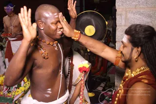 A photograph of a shaved-headed, bare-chested man with rudraksha malas and a white dhoti, smiling with both palms raised, while a man on the right with long swept-back hair, a gold ornamental necklace, a red-and-gold shawl, gold earrings and an orange beaded bracelet touches his forehead with his hand. A standing fan and yellow-and-orange flower garlands sit nearby, and a shirtless attendant in a red-bordered dhoti carries a tray in the background. The right-hand figure has long swept-back hair and distinctive gold adornment, so he is a person present in person; the central man is a devotee receiving blessing.