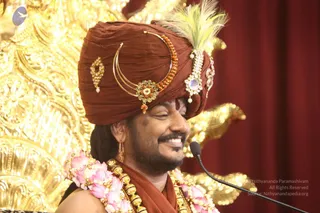A smiling man wears a maroon turban decorated with gold and orange-bead ornaments, a crescent-shaped gold ornament, jewelled brooches and a white-and-green feather plume. He has a shaped beard and a bindi-style mark on his forehead. He wears pink flower garlands and a maroon shawl over his shoulders, and gold beaded adornments hang at his chest. He speaks into a microphone in front of a gold ornate backdrop and red drape.