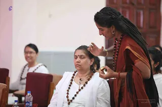 A photograph of a seated woman with closed eyes, wearing a white kurta and a long rudraksha mala, receiving a blessing from a standing woman in a rust-red sari with a gold border. The standing woman has long dreadlocked hair, rudraksha malas, hoop earrings and a bangle, and holds a small gold object in one hand while her other hand is raised over the seated woman's head. Behind them a woman in white with glasses smiles, a water bottle sits on the table and maroon chairs are visible in an indoor hall. Three people are visible in the scene.