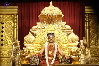 A photograph of a seated man in a golden ornate throne with a cobra-hood canopy and lion-head armrests, set against a deep red curtain. He wears rust-orange robes, a striped headband, a gold necklace, a white and pink flower garland, and sits with eyes closed and hands resting on leopard-print cushions. Swamiji (probable): long dark hair beneath a striped head wrap, rudraksha-style beaded adornments and ceremonial gold necklaces, seated on a throne in darshan.