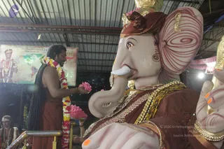 Close view of a large Ganesha murti with a pale pink face, gold crown, red-and-gold dhoti-style drapery and heavy gold necklaces, its trunk and raised hand adorned with gold bangles. In the left foreground a man with long dark hair and a red-saffron cloth wrapped around his body, wearing a garland of white and yellow flowers, offers pink flower petals from a silver tray. Behind him a banner shows a figure in red with a trident and garland beside a map, and an elderly man in white sits at the lower left. Metal railings and a corrugated roof frame the indoor setting, lit by warm lights.