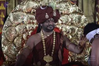 A man seated before an ornate golden halo-shaped backrest with fan-like petals, wearing a maroon turban with a crescent moon ornament and a feather, jewelled brooch pins, and gold bead necklaces with a large gold pendant. He is bare-shouldered with a maroon wrap draped over one shoulder, gold armlets and bangles, and smiles while raising his right hand toward a person in a white head cloth at the right edge.