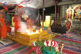 A photograph of a homa fire ritual in a temple-style open hall at night. A fire burns in a square golden fire pit with smoke rising. Several saffron-robed sannyasis in orange and red clothing stand around it; one holds a bowl and flowers, another in an orange turban stands at the fire. Garlands of red and white flowers and brass vessels sit on the striped floor cloth. On the right a figure in orange clothing with a gold necklace sits on a red seat before a microphone stand. Behind them, a shrine with a deity murti and decorated arches, and a dark stone statue with flower garlands.