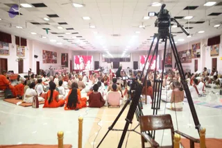 A large hall with white floor and ceiling lights shows a seated congregation of many people, most in orange, saffron and white robes, sitting cross-legged on mats. In the foreground a professional video camera on a tripod records the gathering. Two large screens at the front display a figure in red and white, and a seated figure in white is visible near the stage. Wall displays and framed images line the room, and a wooden chair stands in the front corner.