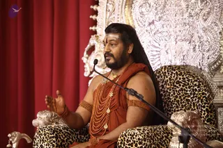 A man seated on a leopard-print upholstered throne speaks into a microphone on a stand. He wears saffron-orange draped clothing, multiple strands of orange rudraksha malas, gold pendant necklaces, and orange beaded armlets, with a white and red forehead mark and long dark hair swept back. He gestures with his right hand open. Behind him is an ornate silver-embossed throne back and red curtains.