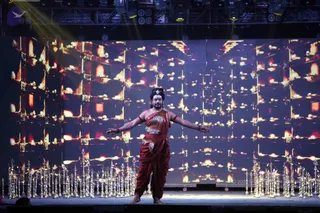 A photograph of a single male performer standing barefoot on a stage with arms spread wide. He wears a deep red draped garment with a cream and black patterned panel across the chest and waist, a red sash-like wrap, and bangles on the upper arms and wrists. A tilak mark and a small ornament adorn his forehead, and his hair is tied up. Behind him a large video wall shows glowing oil lamp imagery in orange and gold. Rows of ornate brass oil lamps stand in front of the stage on both sides. Stage lights hang overhead. The figure's appearance does not clearly establish an identity, so no candidate is named.
